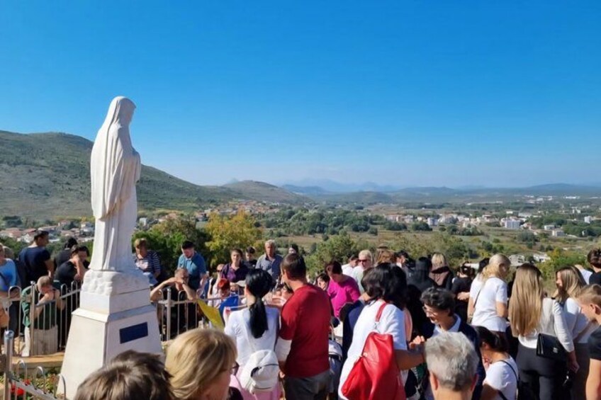 A crowd gathered around a statue on a hill with a view of a town and hills in the distance. 3