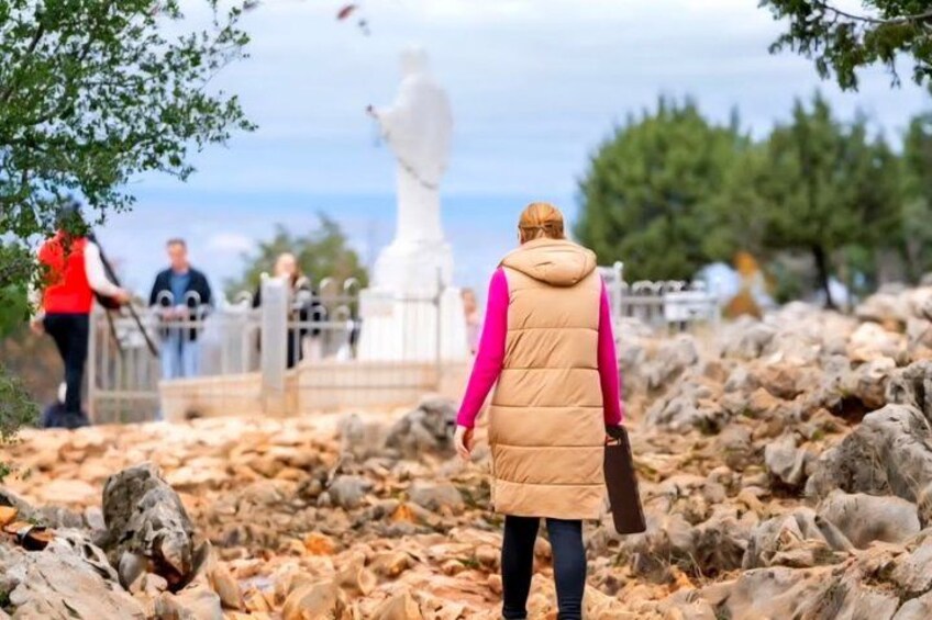 A person in a pink jacket and beige vest walking on a rocky path with a statue in the background. 2