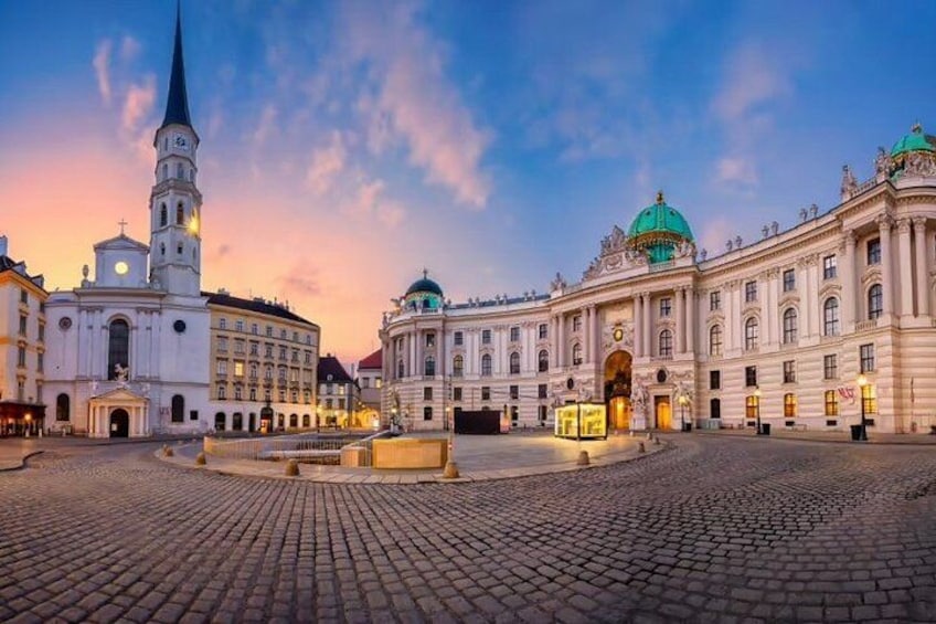 A historic square with cobblestone pavement, a church with a tall spire, and a grand building with a green dome. 1