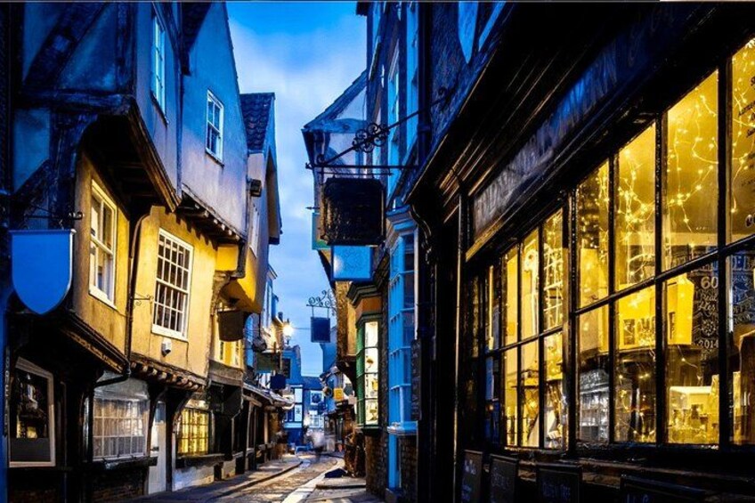 A narrow street with old buildings, illuminated windows, and a cobblestone path. 5