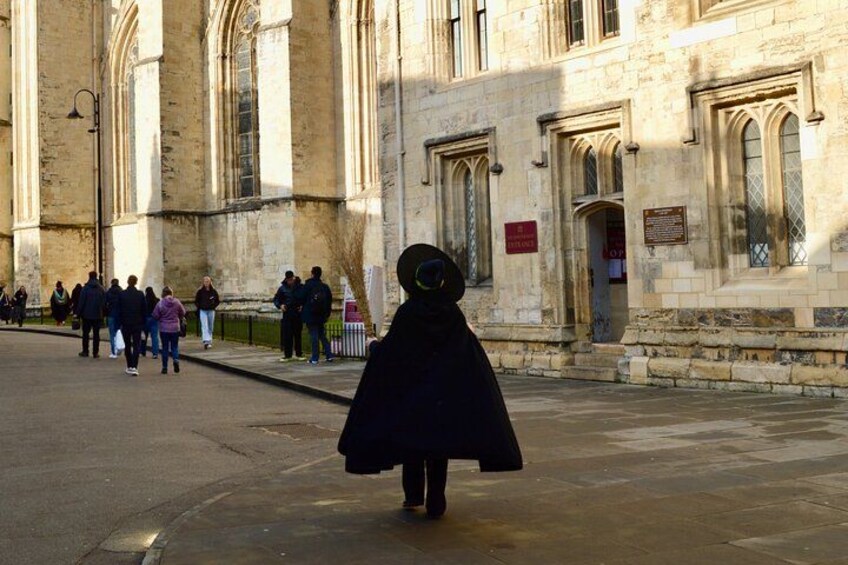 A person in a black coat walking on a street in front of a historic building with arched windows and stone walls. 2