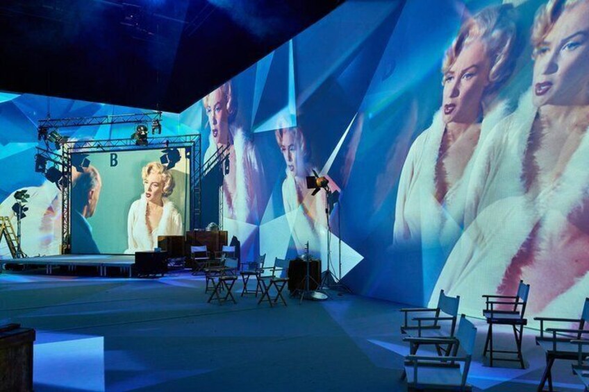 A stage with a large screen displaying a person in a white outfit, stage lighting, and a backdrop with geometric patterns. 1