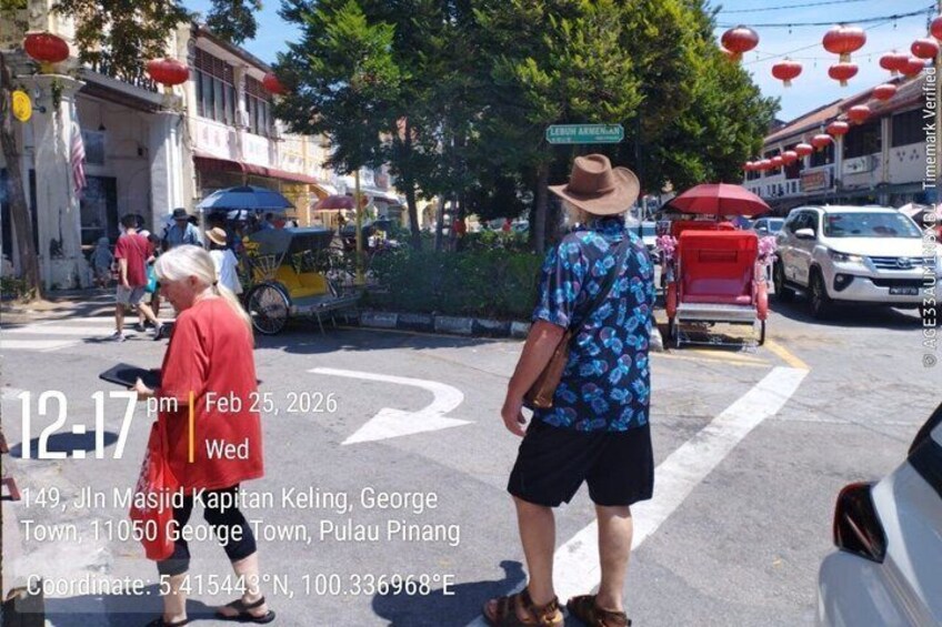 Una calle con un hombre de camisa y sombrero azul, una mujer con un vestido rojo y un tuk-tuk. 2