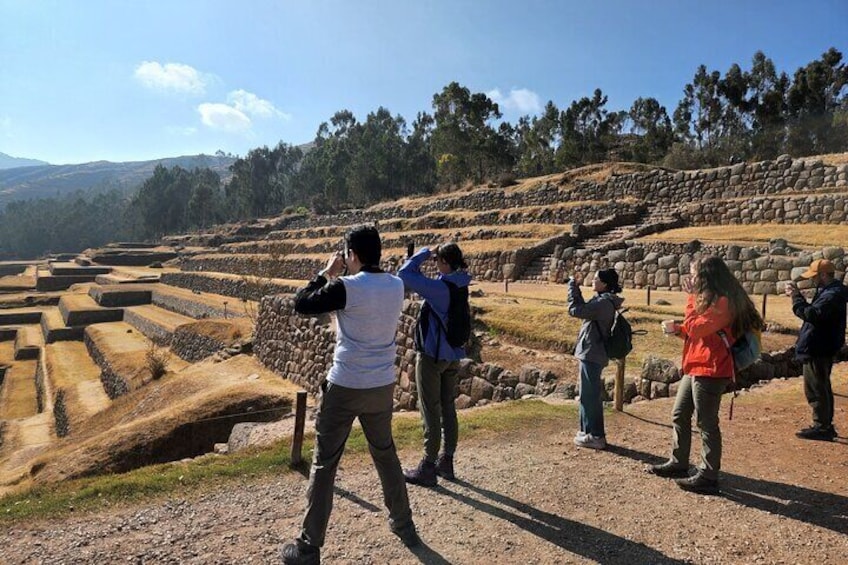 Tourists taking photos at an archaeological site with terraced structures and a mountainous backdrop. 1
