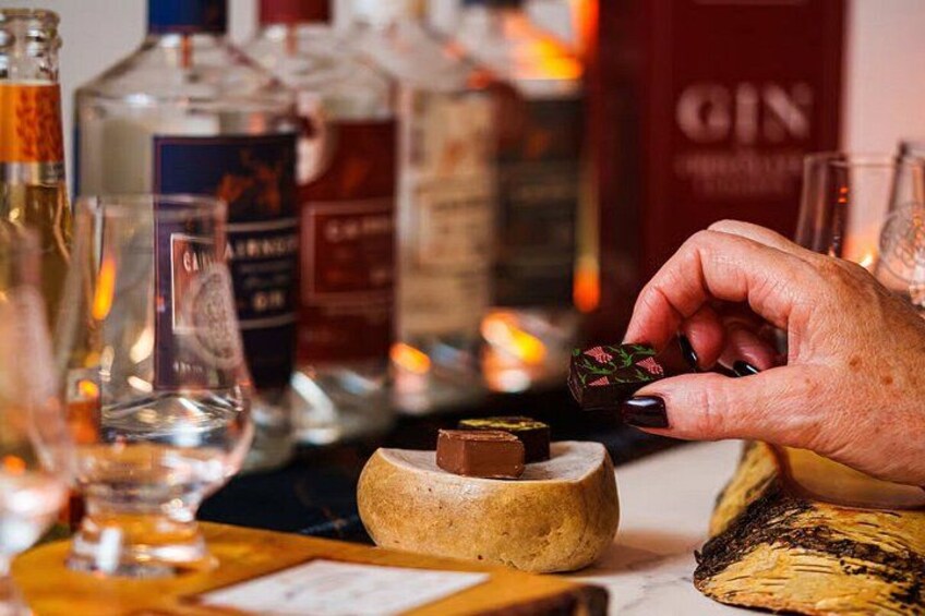 A hand holding a chocolate piece over a small bowl, with various bottles and glasses in the background. 3
