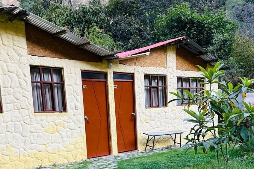 A stone building with two brown doors and windows, a small table, and a grassy area with plants. 4