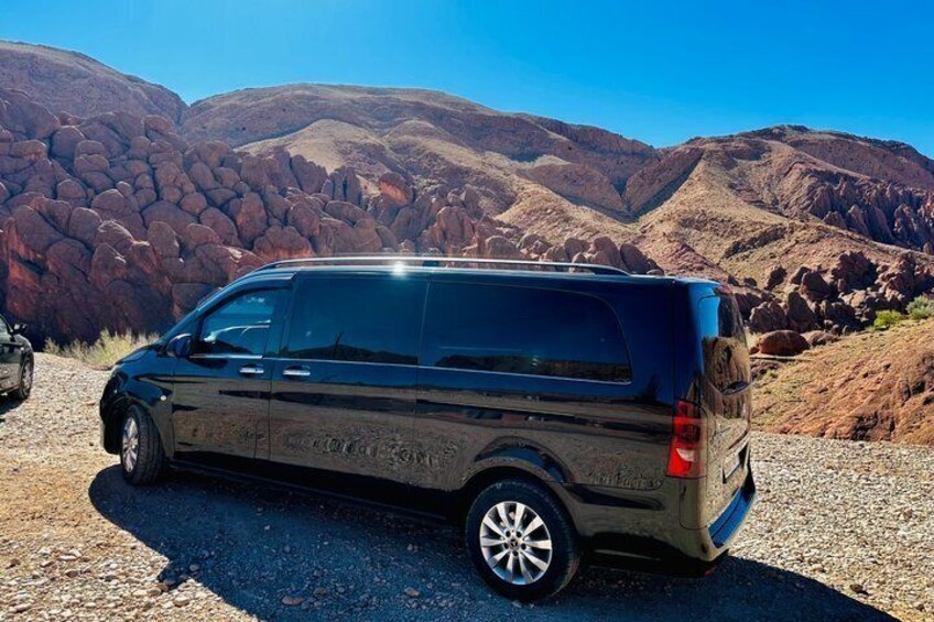 A dark-colored minivan parked on a gravel road with red rock formations in the background. 3
