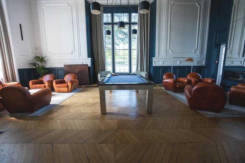 A billiards table in a room with wooden flooring, surrounded by leather chairs, and a fireplace. 3