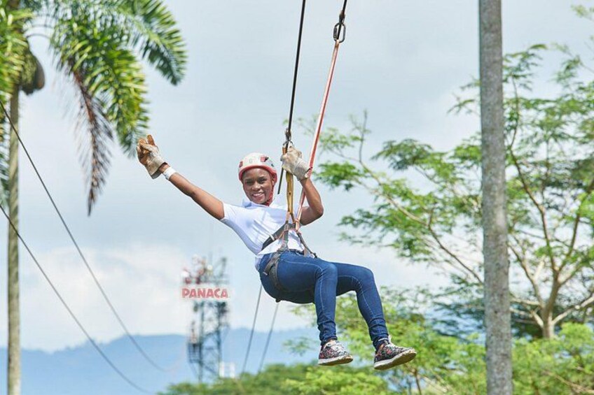 A person zip-lining with a harness, wearing a white t-shirt, blue jeans, and sneakers, with a palm tree and a sign reading 'PANACA' in the background. 3