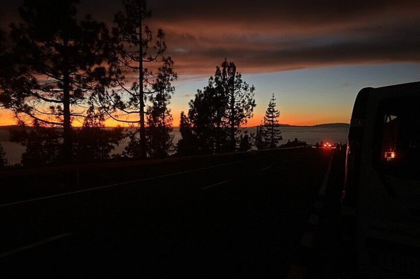 A sunset with trees silhouetted against the sky, a road, and a vehicle's rear lights. 4