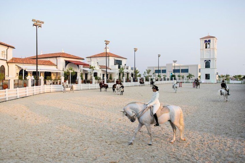 A horse riding arena with riders and horses, surrounded by buildings and a clear sky. 2