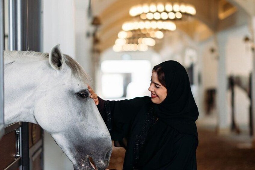 A person petting a white horse inside a stable with wooden stalls and a chandelier in the background. 1