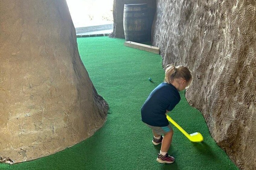 A person playing golf indoors with a yellow club, green turf, and artificial rock walls. 1