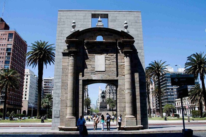 A large stone archway with people walking around it, surrounded by palm trees and modern buildings. 3