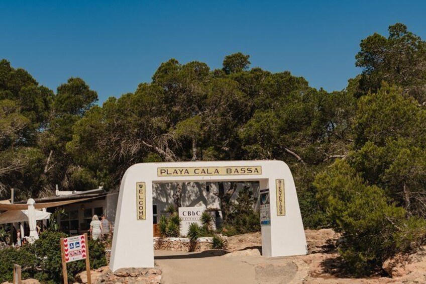 A white archway with 'PLAYA CALA BASSA' written on it, surrounded by trees and a building in the background. 5
