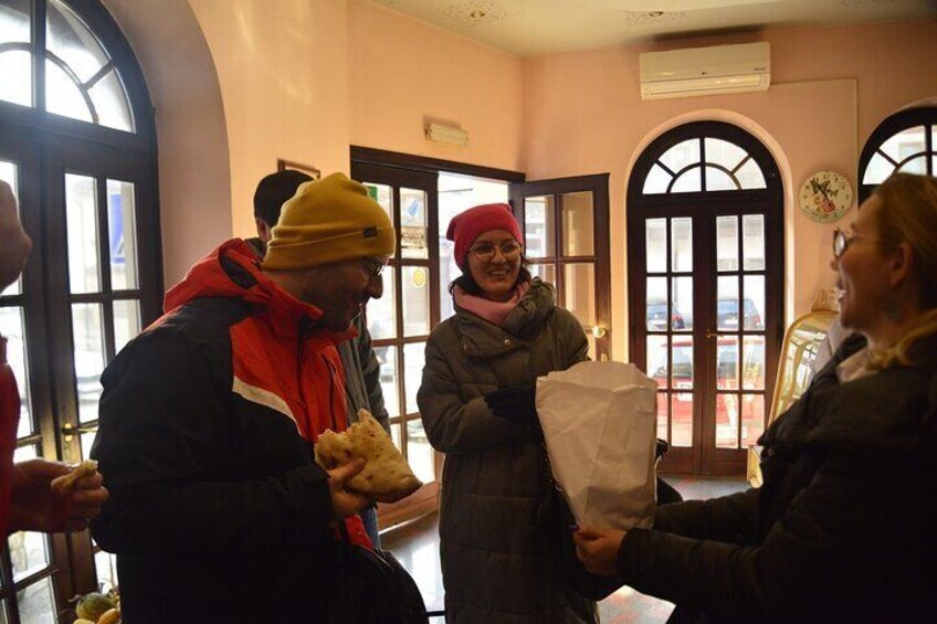 A group of people indoors, some holding food items, with arched windows and a door in the background. 3
