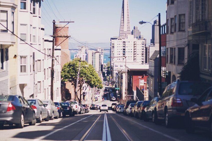 A city street with parked cars, buildings, and a prominent tower in the distance. 1