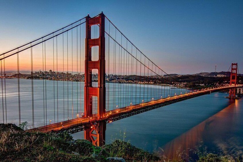 A suspension bridge with towers, a calm body of water, and a city skyline in the background. 3