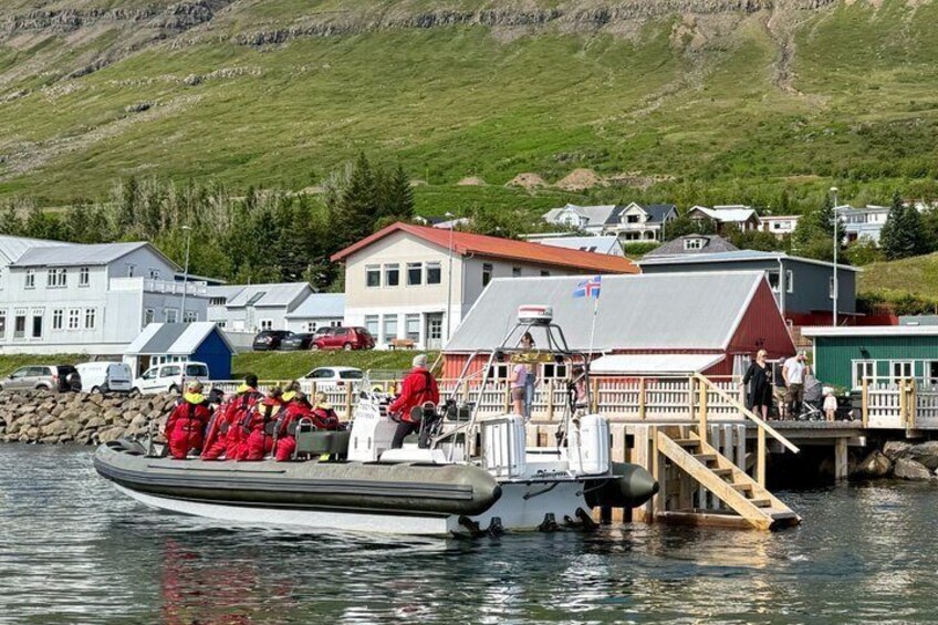 A boat with people in red suits docked at a pier with houses and a mountainous backdrop. 2