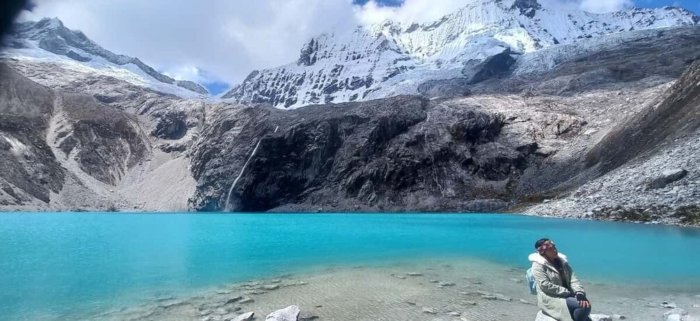 A person standing by a turquoise lake with snow-capped mountains in the background. 1
