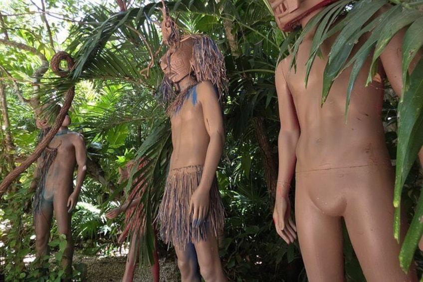 A sculpture of a person with a headdress and a skirt made of natural materials, set in a tropical environment with dense foliage. 5