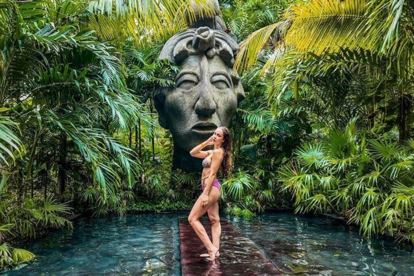 A person in a bikini standing in a water feature surrounded by lush tropical vegetation and a large stone sculpture. 1