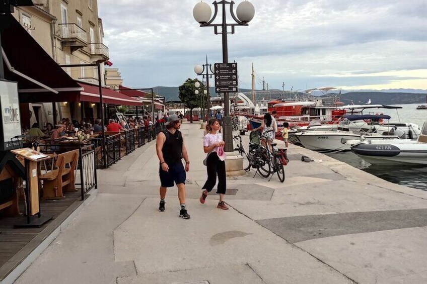 A waterfront promenade with people walking, a café, and boats docked at the marina. 2