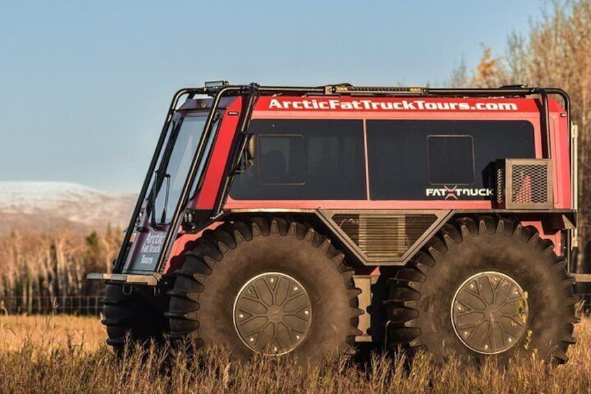 A large red truck with 'ArcticFatTruckTours.com' and 'FAT TRUCK' written on it, parked in a field with mountains in the background. 3