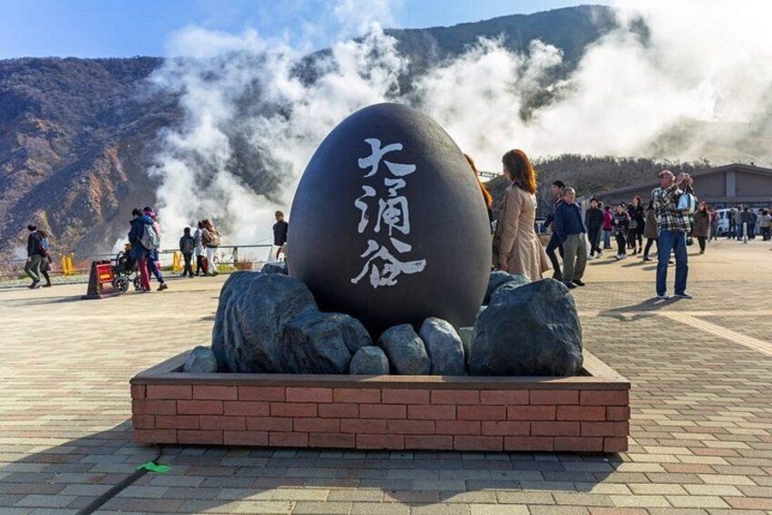 A large stone sphere with Japanese characters, surrounded by rocks and a brick base, with people walking around and steam rising in the background. 4