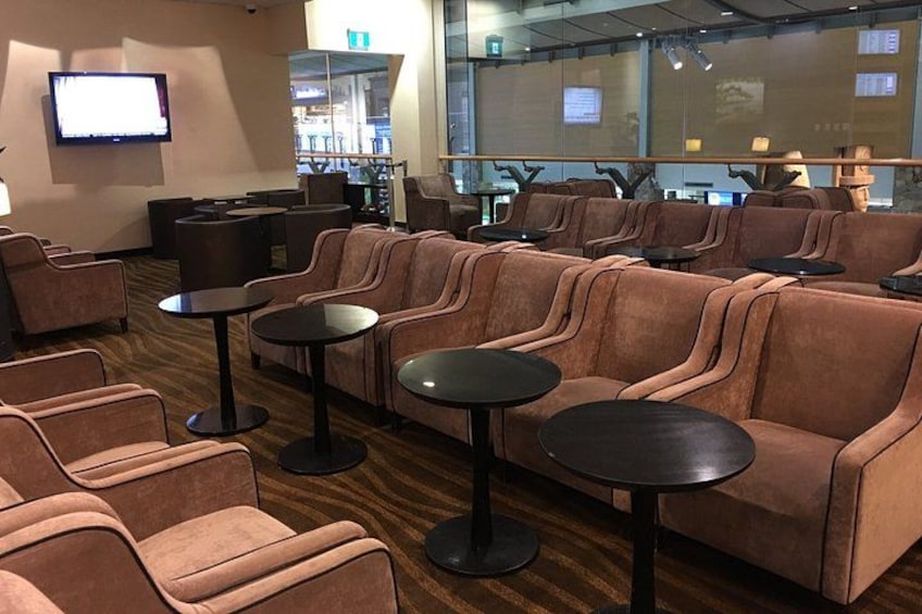 A lounge area with sofas and small round tables, a television screen, and a glass partition with a view. 4