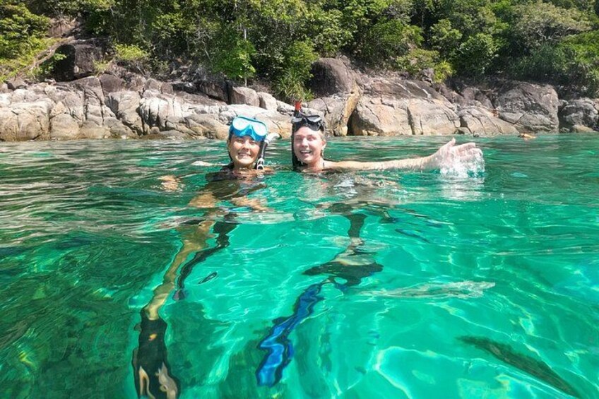 Twee mensen snorkelen in helder turquoise water, met een rotsachtige kustlijn en groen in de achtergrond. 5