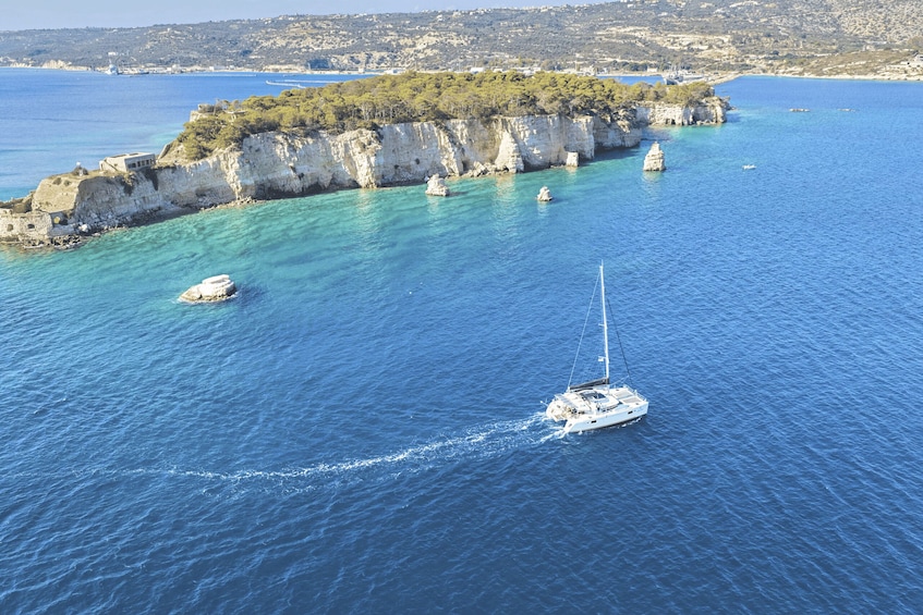 A sailboat on a clear blue sea with rocky cliffs and a small town in the background. 1