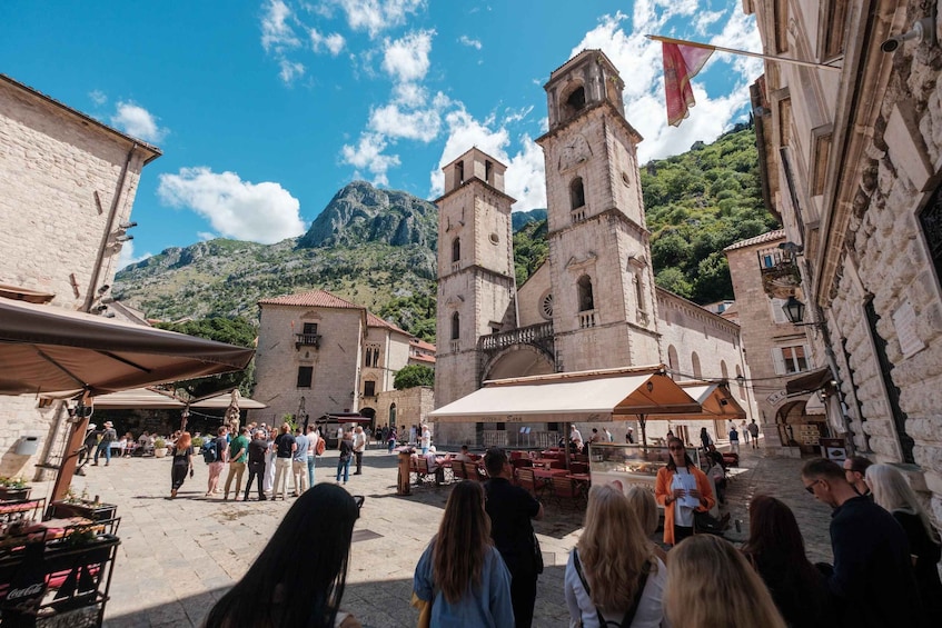 A historic square with stone buildings, a clock tower, and a mountainous backdrop. 2