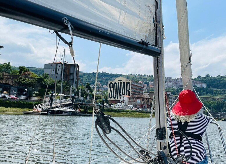A sailboat with a person wearing a red hat and striped shirt, ropes, and a cityscape with a sign reading "SONAR" in the background. 5