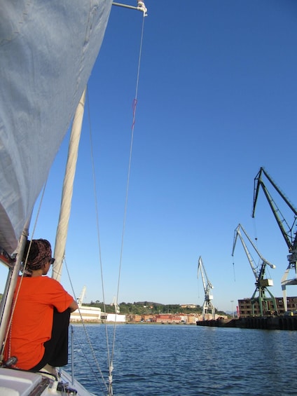 A sailboat with a person in an orange shirt, a clear blue sky, and industrial cranes in the background. 2