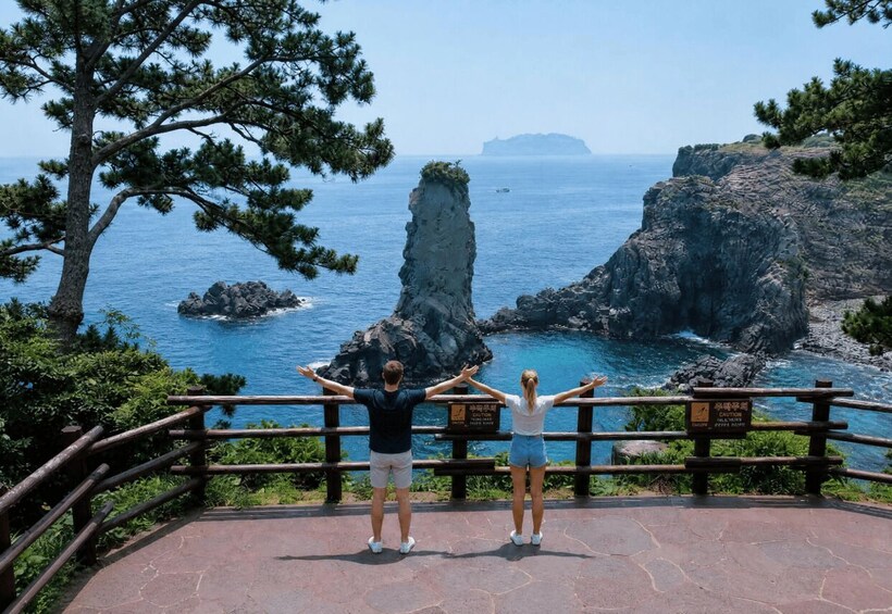 Two people standing on a viewing platform with a railing, overlooking a coastal landscape with rocky cliffs and a clear blue sea. 5