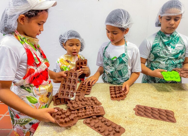 Children wearing hairnets and aprons, engaged in making chocolate shapes with molds. 2