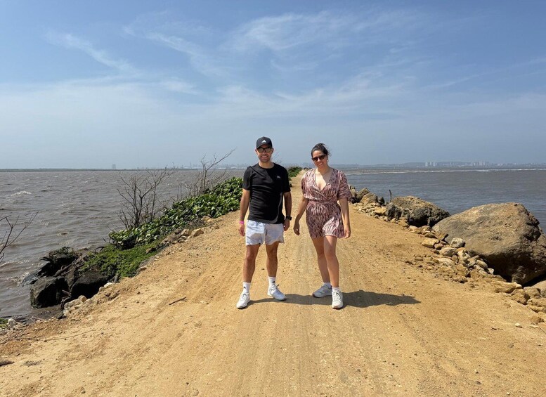 Two people standing on a dirt path by a body of water with rocks and vegetation. 1