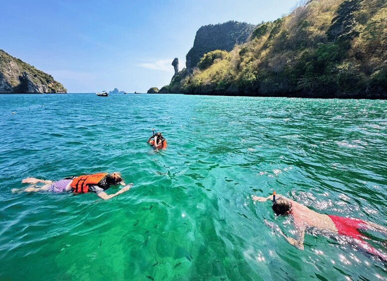 Three people swimming in clear turquoise water with a rocky coastline and cliffs in the background. 5