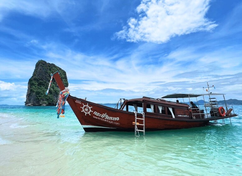 A traditional wooden boat with a ladder, anchored near a clear beach with a large rock formation in the background. 3