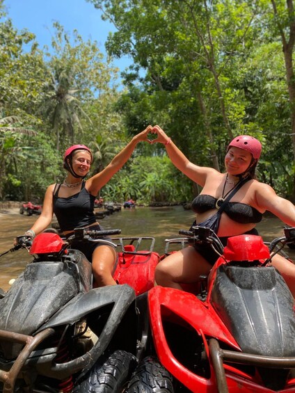 Two people on red all-terrain vehicles in a forested area with a river. 3
