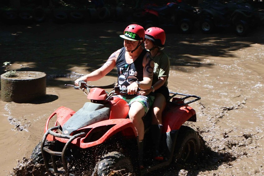 Two people on a red ATV in a muddy area with a water body in the background. 5