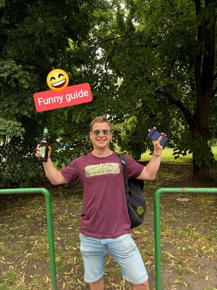 A person holding a bottle and a smartphone, standing in a park with green railings and trees. 2