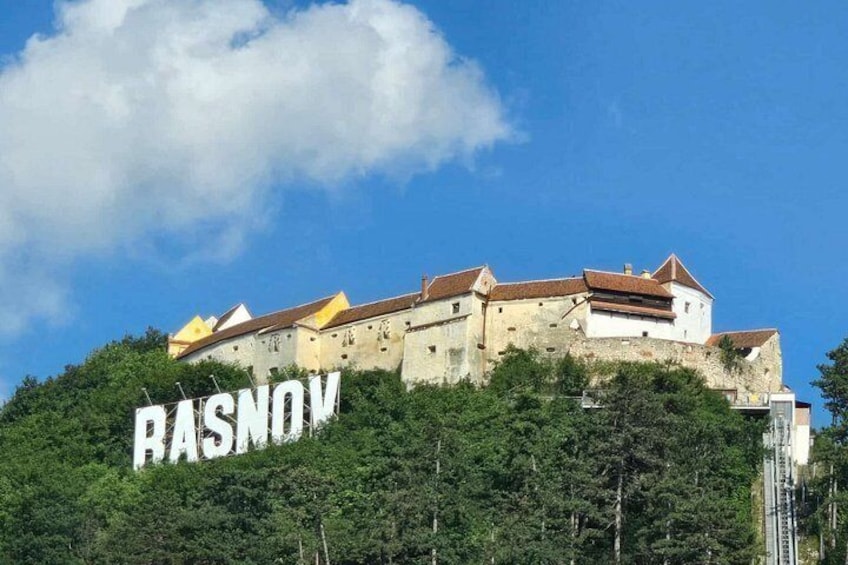 A historic building with the word 'RASNOV' on its facade, surrounded by lush greenery. 5