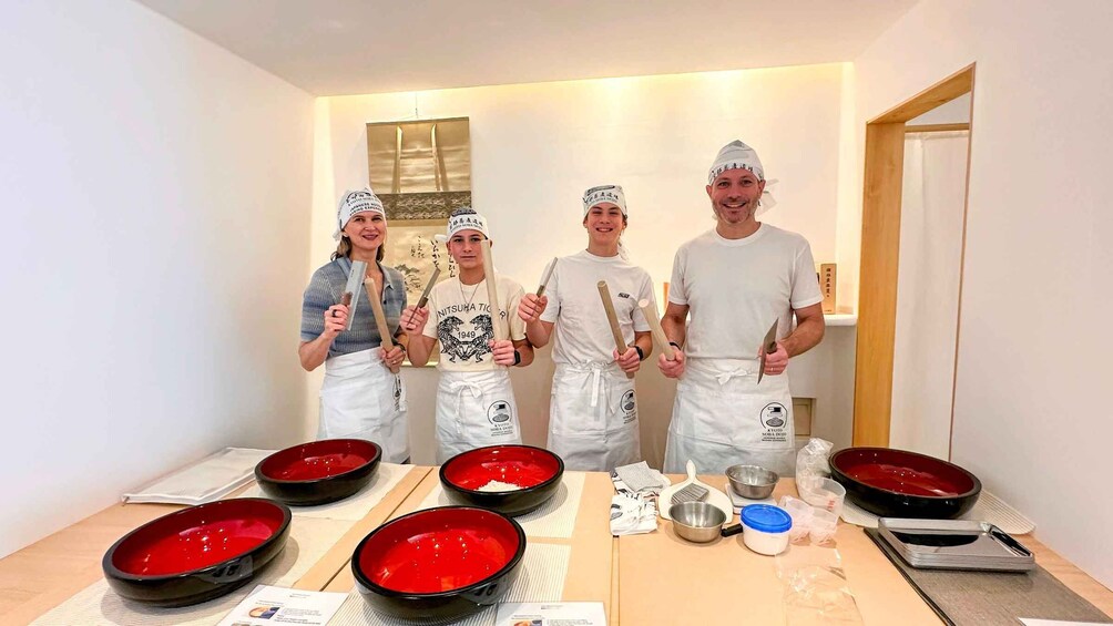 Four people in a kitchen holding wooden utensils, with red bowls on a table. 1