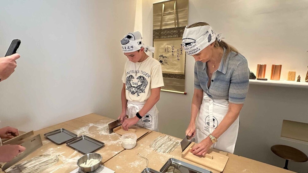 Two people preparing food, one cutting dough and the other rolling it, with a table covered in flour and kitchen tools. 4
