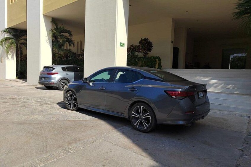 A gray sedan parked in a covered parking area with palm trees and a building in the background. 2