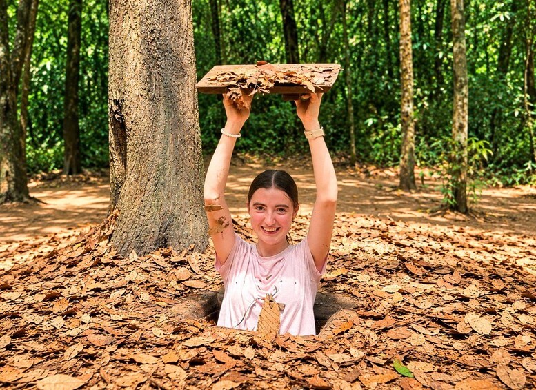 A person holding a wooden board above their head in a forest with a tree trunk and fallen leaves on the ground. 1