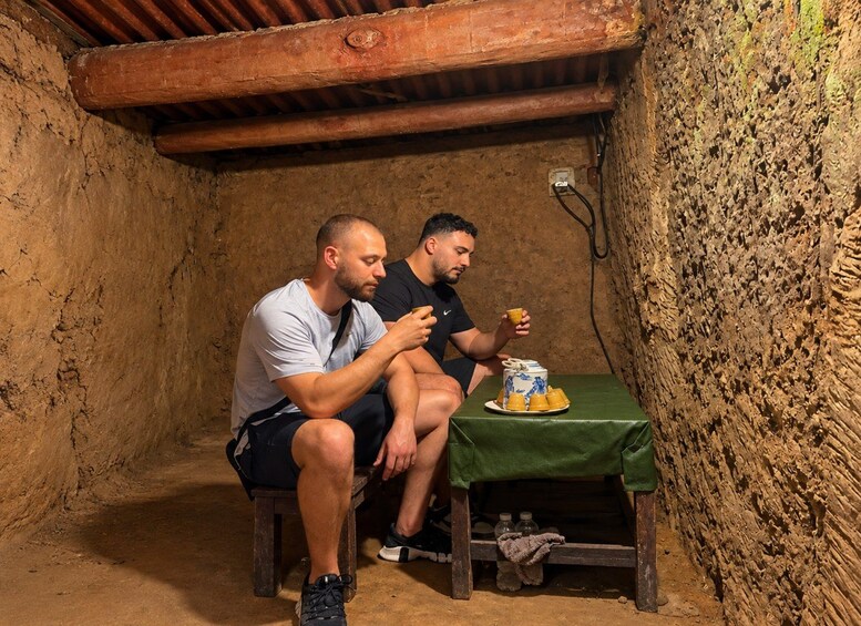 Two men sitting on stools in a rustic stone-walled room with a wooden ceiling, a table with a cake and cups, and a power outlet on the wall. 5