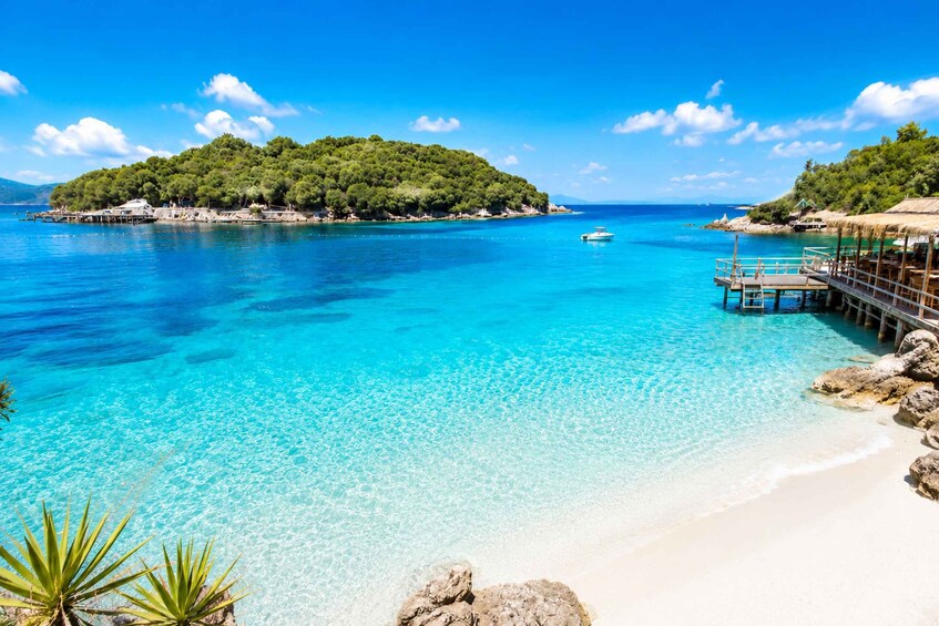 A turquoise sea, a sandy beach, a wooden pier, and a forested island. 1